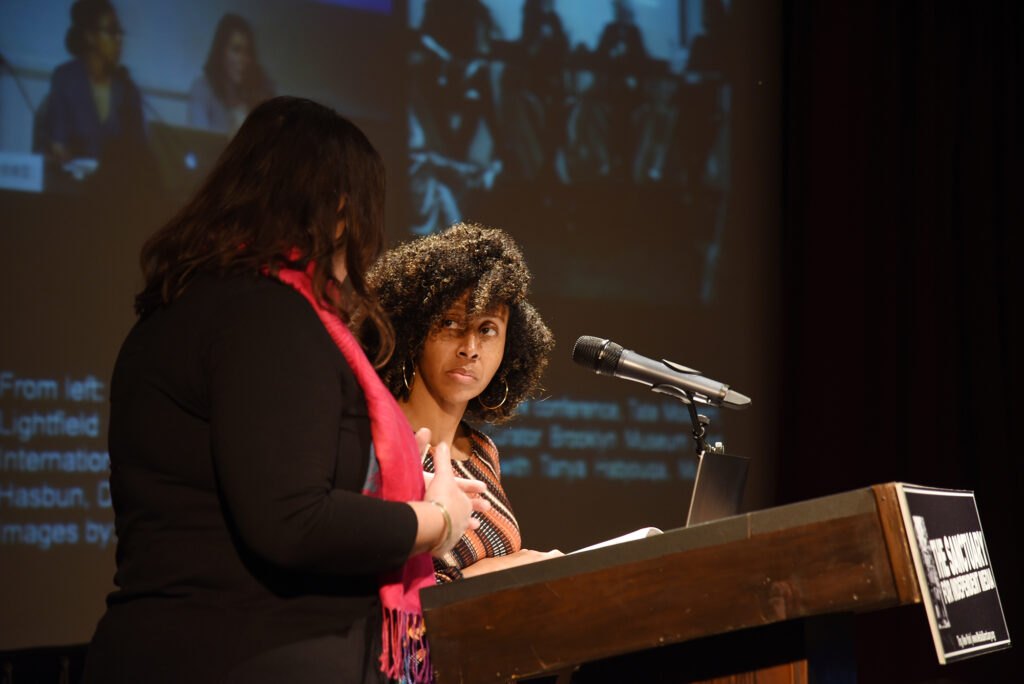Lesly Deschler Canossi and Zoraida Lopez-Diago of Women Picturing Revolution stand behind the podium at The Sanctuary. An image from their book, 'Black Matrilineal, Photography, and Representation' is projected on the screen behind them.