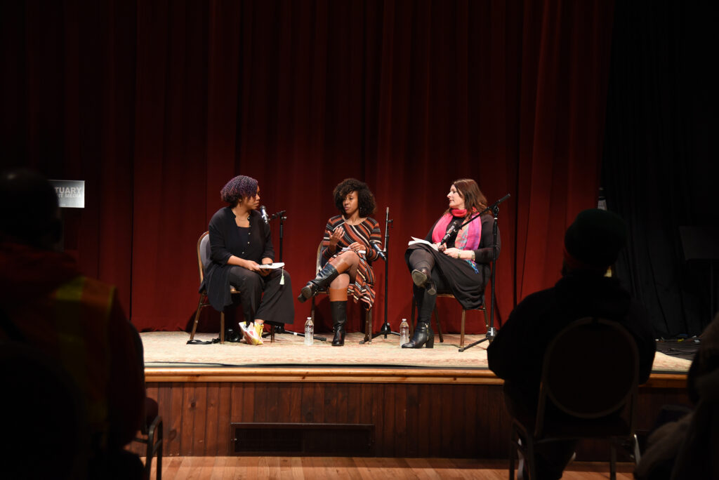 Jayana LaFountaine, Zoraida Lopez-Diago, and Lesly Deschler Canossi sit on stage at The Sanctuary for Independent Media with microphones with a red stage curtain in the background. Audience members can be seen in silhouette in the left and right foreground.