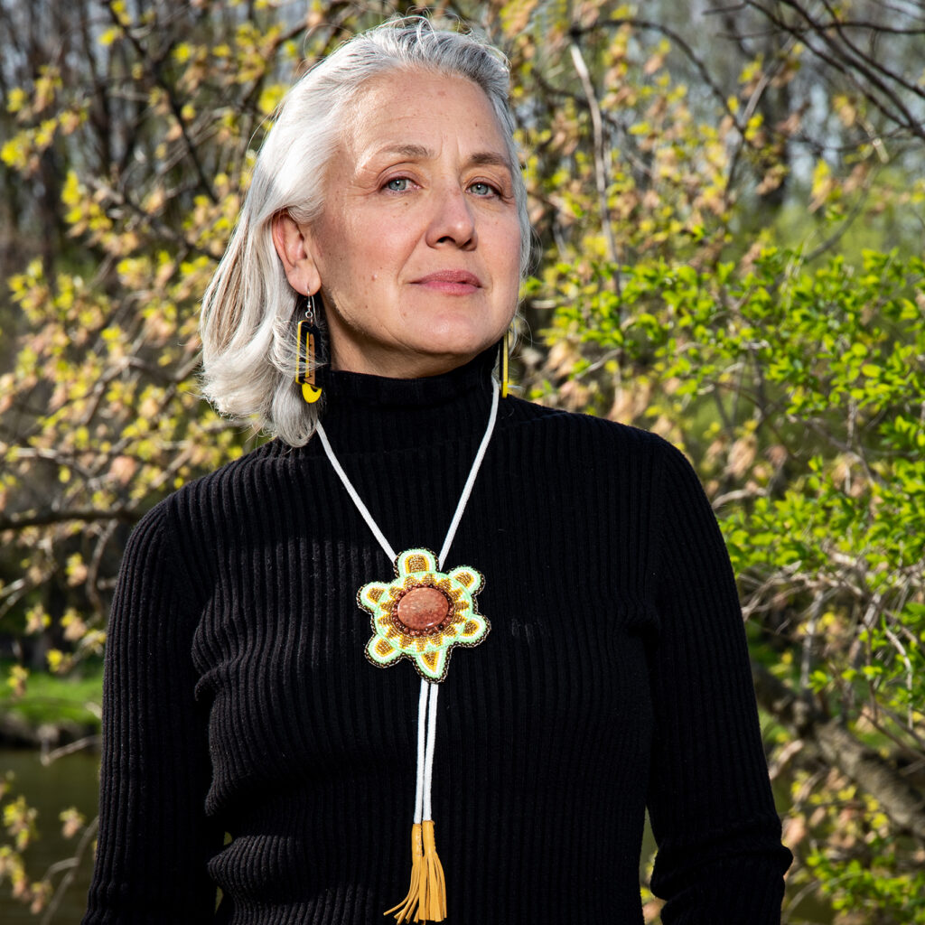 Headshot of Kahstoserakwathe Paulette Moore outside in front of trees. She wears a black turtleneck top, dangling earrings, and a turtle clan necklace. She has white hair to her shoulders.