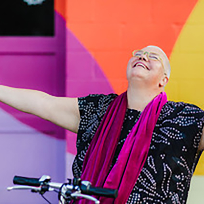 Petra Kuppers 21: Petra Kuppers, a white queer disabled cis woman of size with yellow glasses, shaved head, pink lipstick and a black dotted top, smiles up to the sky, arms outstretched, embracing the world. Her mobility scooter’s handlebar is visible at the bottom of the image. She is in front of a multicolored wall: purple, pink, yellow and orange.