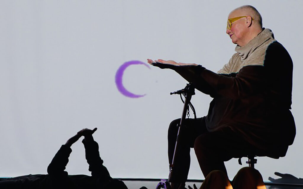 Starship Somatics moon: Petra, a white queer disabled cis woman of size sits on her scooter, and reaches out her hand to a Starship Somatics video projection of a purple moon. Below, a floor dancer reaches up and touches their own hands. Photo: video still, Petra Kuppers