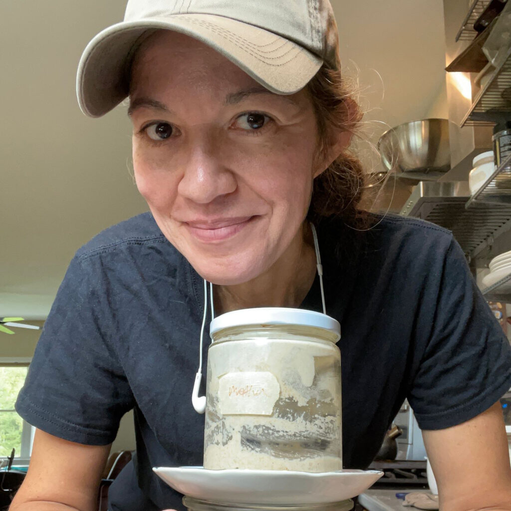Ellie Markovitch leans over jars with cooking material for a picture. She has a beige cap and has earbud headphones around her neck