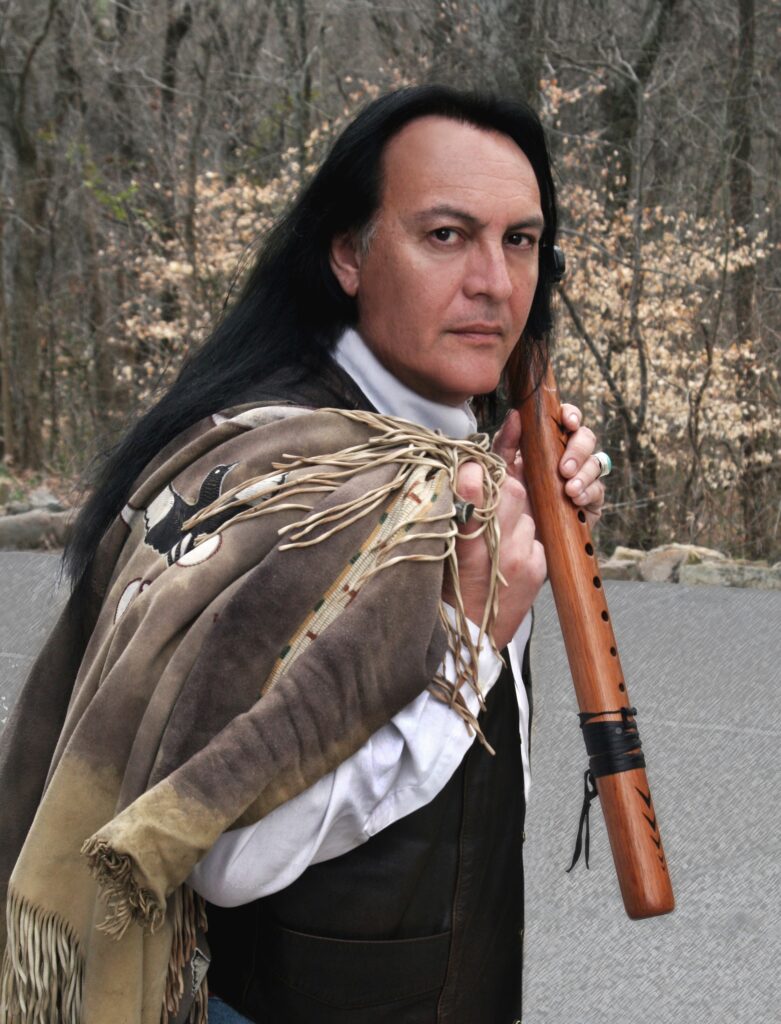 Bill Miller, an Indigenous man, has a leather jacket over his shoulder and is holding a wooden flute. He is wearing a while short and black vest and has long, black hair. He is standing by trees.