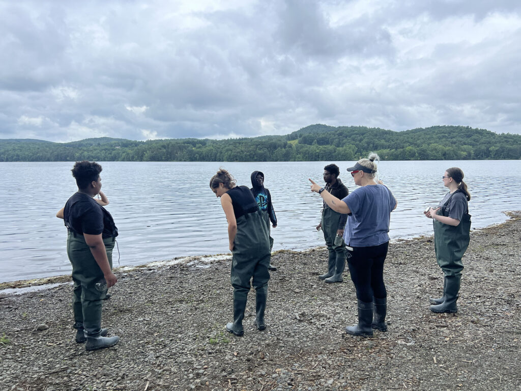 Sarah stands on the pebbled shore of a body of water with a group of Sanctuary youth fellows. Sarah gestures broadly with her hands, apparently talking. 