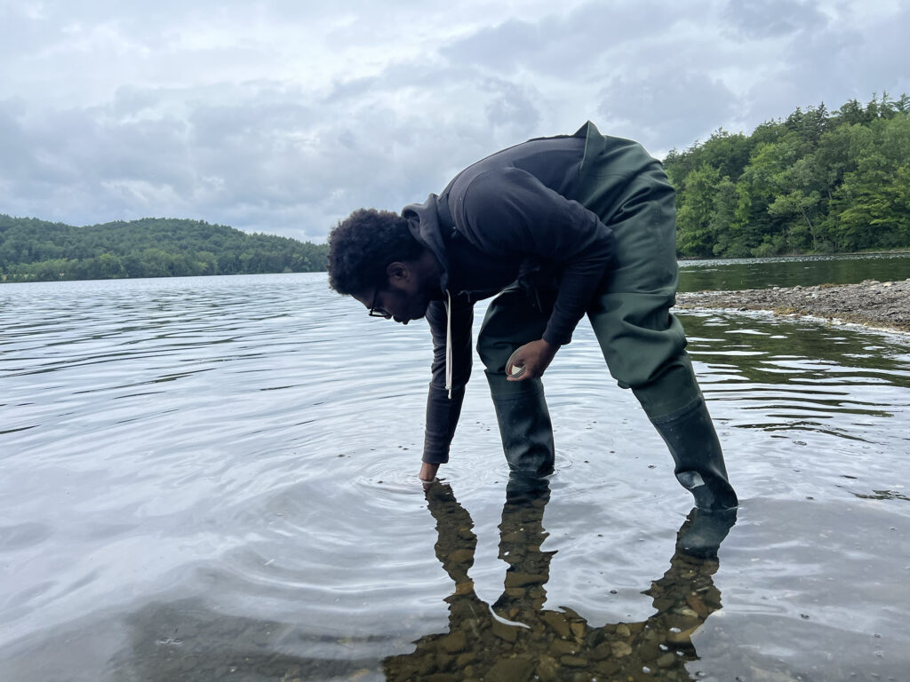 A dark-skinned young man, wearing waders, stands in ankle-deep water and reaches to the bottom, creating ripples. 
