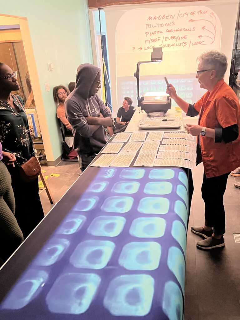 Kathy High leads a presentation on water contamination in the NATURE Lab. An oversized printout of water sample cells rests on the metal lab table. 