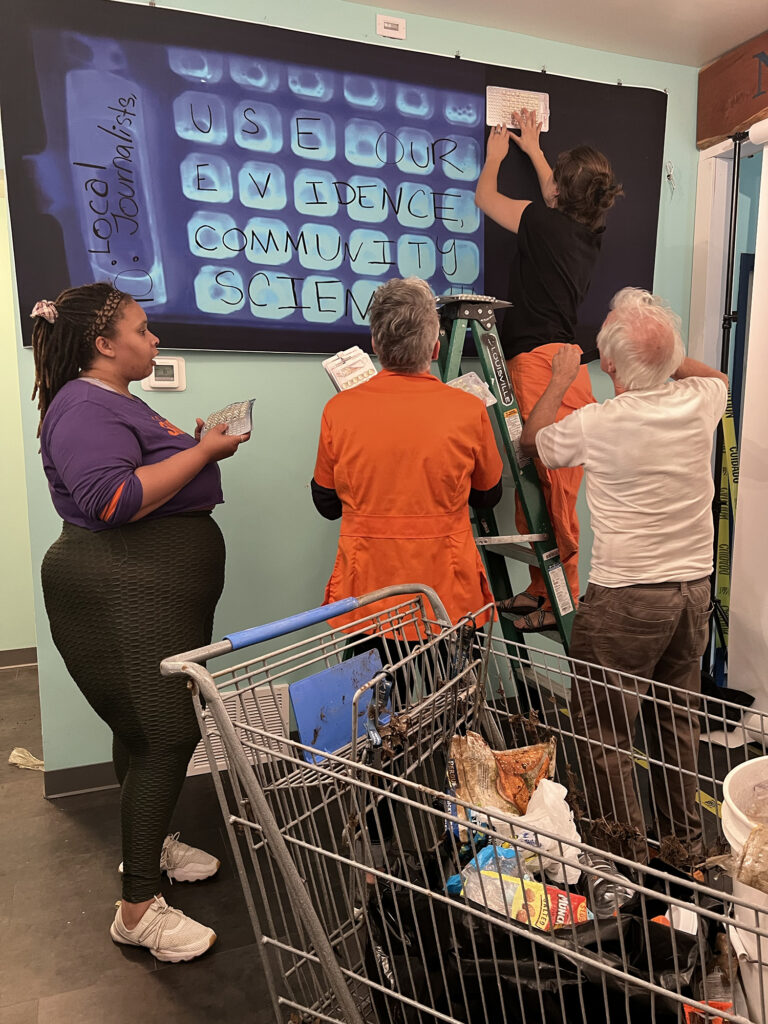 Four people examine a projection of water sample cells and take notes on sheets of paper. In the foreground is a shopping cart containing an open bag of litter. 