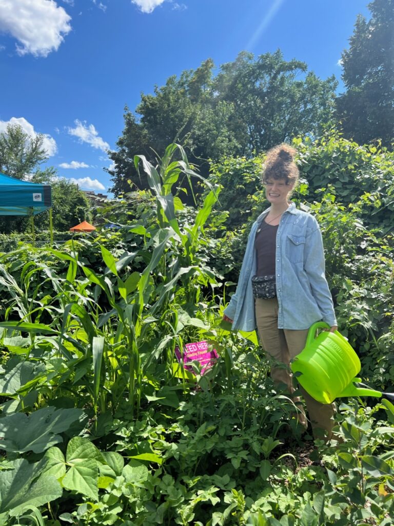 Sienna Thiem is pictured in the lush Collard City Growers Garden wearing a blue long sleeve and khaki pants. She is holding a green watering can among the various plants in the garden on a bright sunny day.