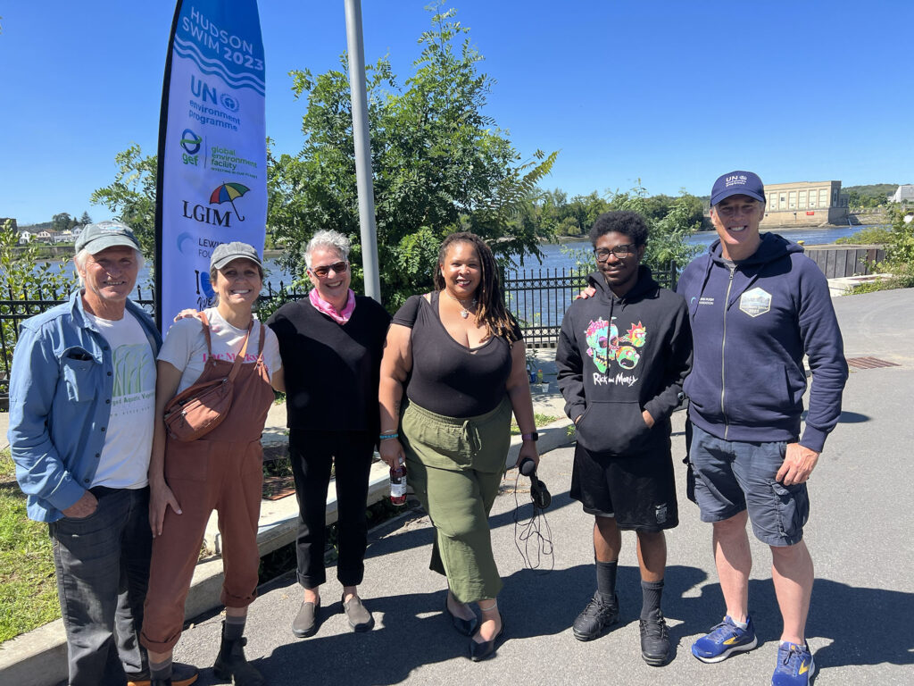 Members of the Water Justice Lab team (5 people of varying ages and races, including L to R: volunteer & mentor Doug Reed, Community Science Educator Ellie Irons, NATURE Lab coordinator Kathy High, Media Mentor Alÿcia Bacon, and Media Intern Aljahraun Wright) pose with Lewis Pugh (far right) at the Ingalls Avenue Boat Launch, with a view of the Hudson River behind them. A banner in the background reads "Hudson Swim 2023".