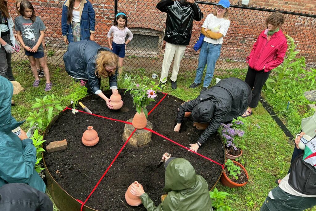 a high-angle photograph of people planting clay vessles and plants into a round, raised garden. In the center is an elevated clay vase with ribbons going outward, partitioning the circle in four.
