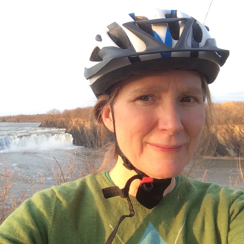 Amy Halloran taking a selfie wearing a bike helmet with a river behind her