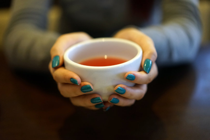 Two hands with turquoise nail polish wrapped around a white cup of reddish colored tea.