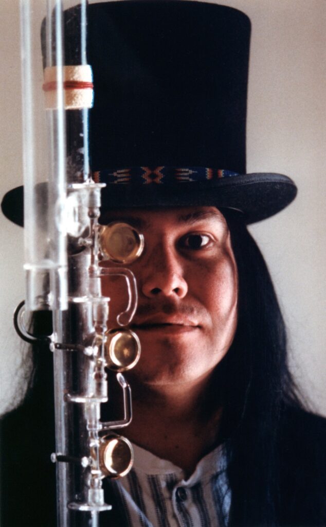 Composer Brent Michael Davids wears a top hat over long, straight, black hair in the background, with a glass flute in front of the left side of his face.