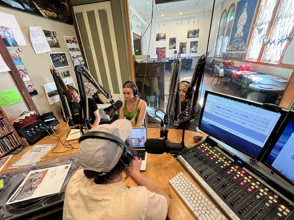 the photo was taken behind the engineer who is sitting with headphones at the soundboard. On the other side of the table are three participants who also wear headphones and are sitting in front of microphones. Behind the three participants is a big glass window behind which we see the Sanctuary cafe with gallery and large windows bringing in daylight to the right