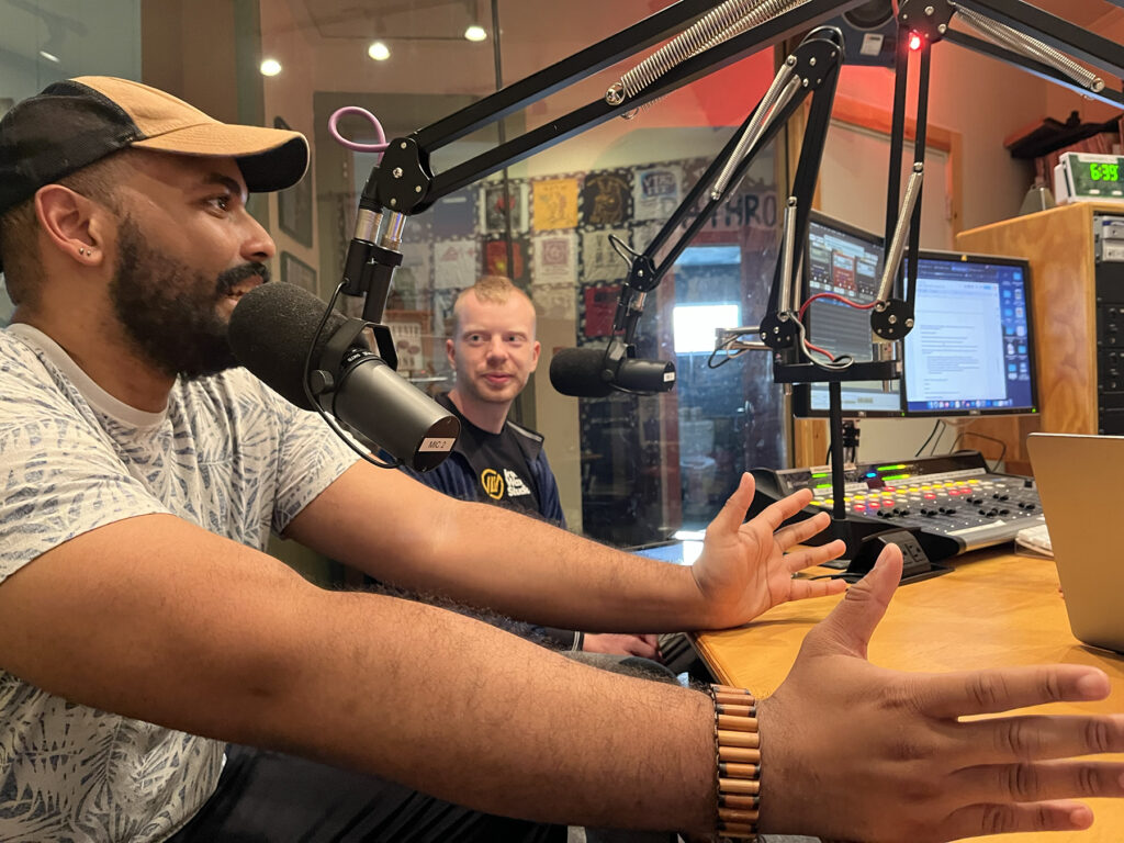 two people are talking into mics. In the foreground is Greg Creft who is speaking into the mic and expressing with his body through outstretched hands. In the background is Brad Monkell who is listening to Greg and also in front of a mic.