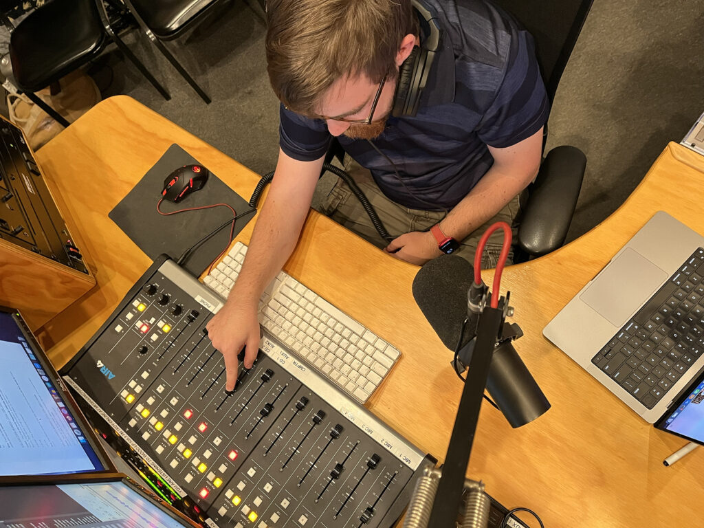 a high angle photo of Caelan McPherson at the soundboard in the radio studio. His finger is moving the fader