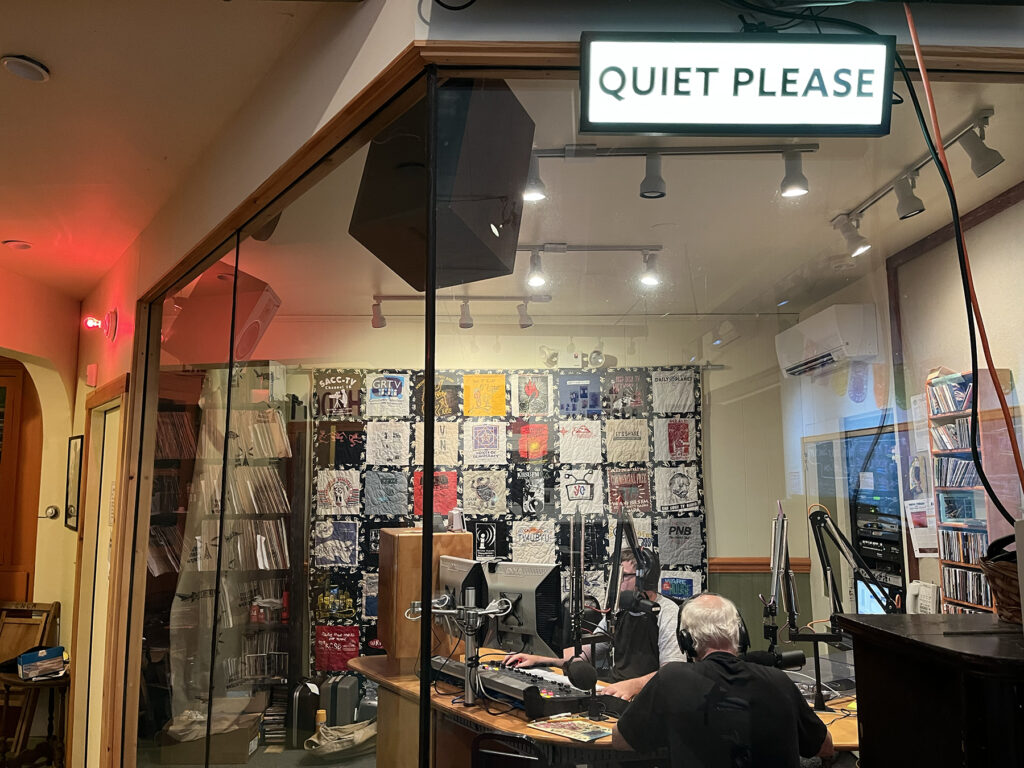 a photo from outside the radio studio. Inside is an engineer and a host on the otherside of a table at a mic. Outside the glass studio walls are signals that radio is on air with the "quiet please" sign alight in white and a red light over the entrance to the studio.