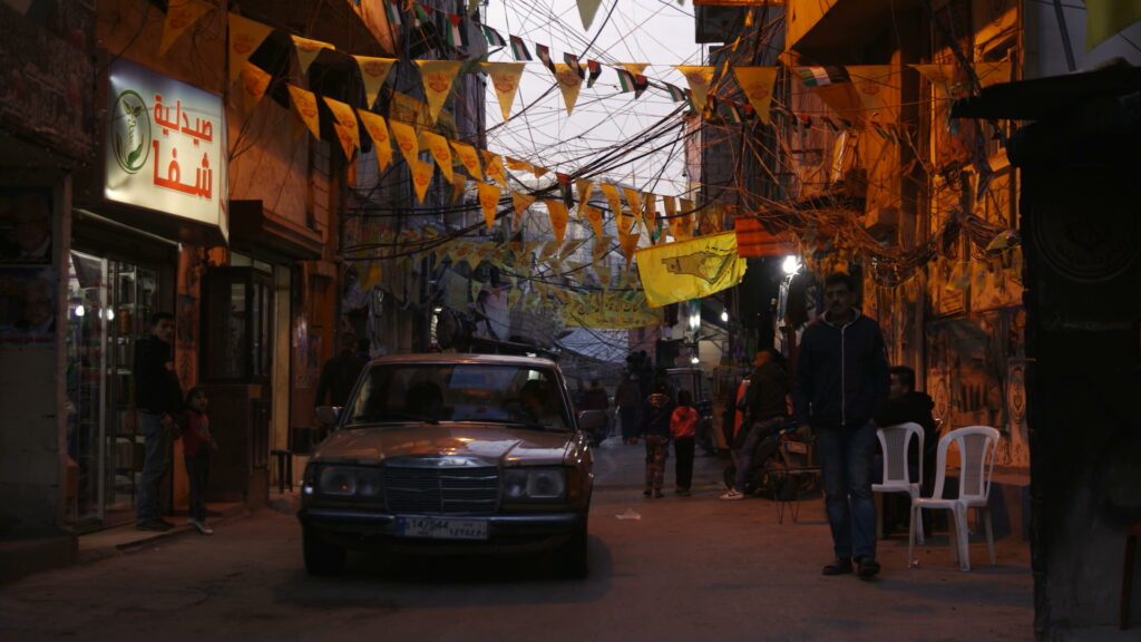 Still from "Spaces of Exception" showing a dark street with yellow banners, lots of wires, a car, and arabic signs