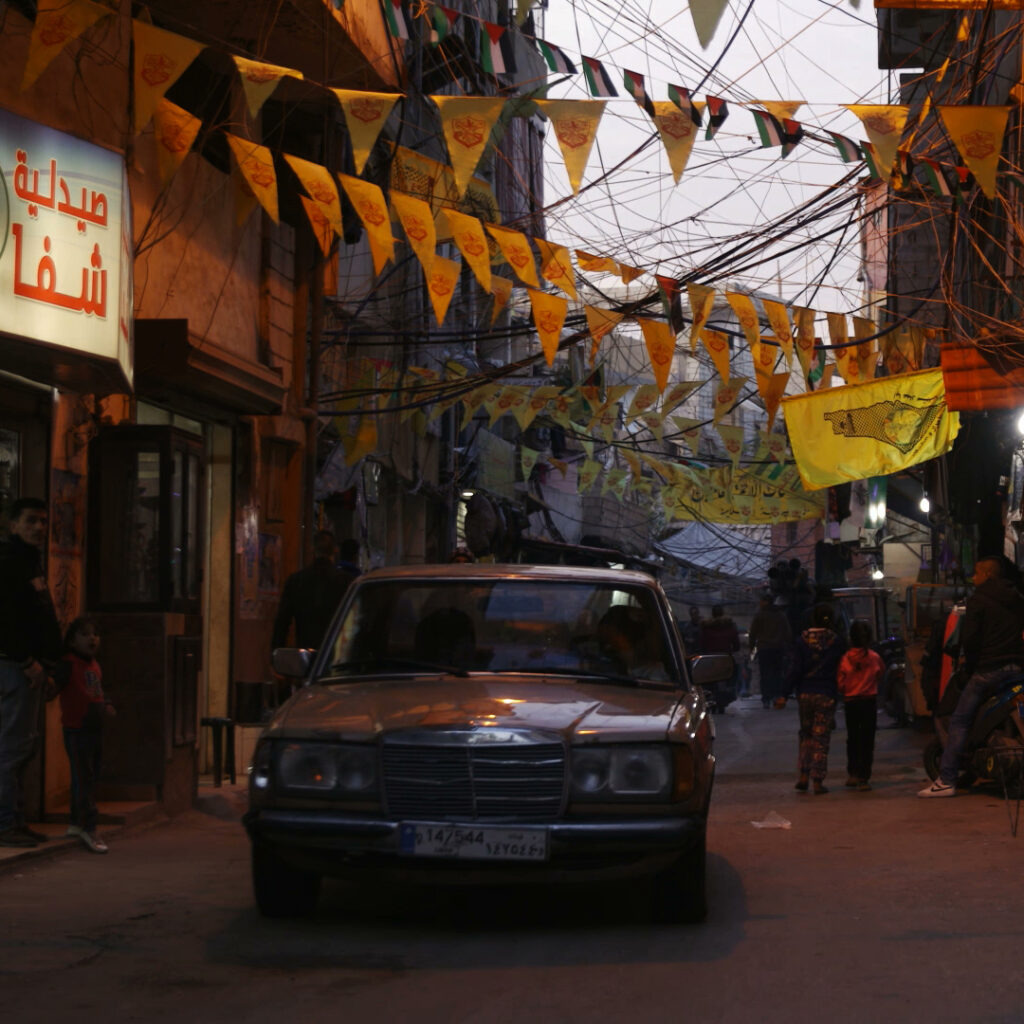 Still from "Spaces of Exception" showing a dark street with yellow banners, lots of wires, a car, and arabic signs