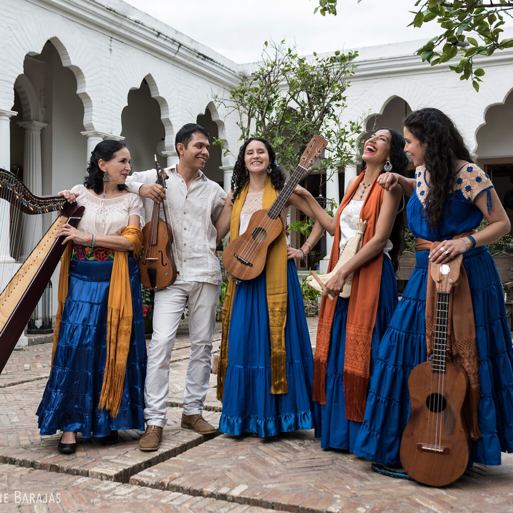 A group photo of Caña Dulce y Caña Brava. They are in a courtyard, dressed in raditional attire of Veracruz with instruments: harp, violin, guitar, an jawbone percussion.