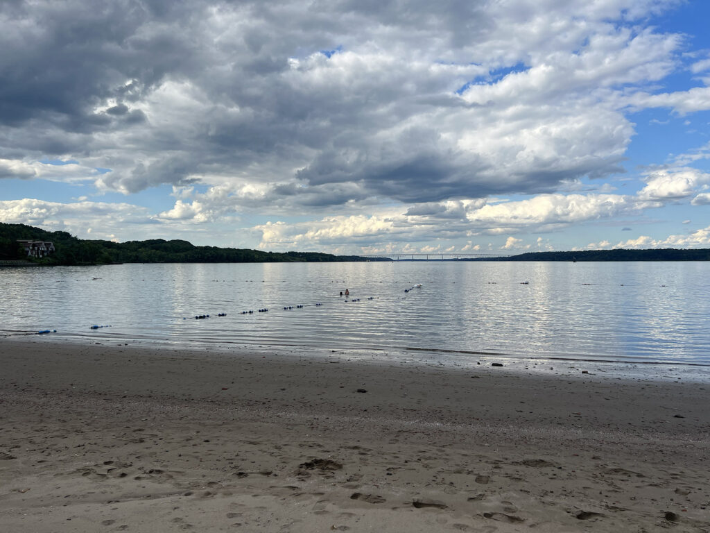 A view of a sandy beach, water, and puffy clouds in the sky, with a shoreline visible in the distance.