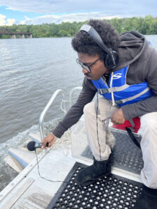 Aljaruan Wright, wearing headphones, leans over the side of a boat recording sound with a microphone