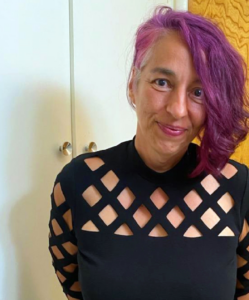 A portrait of a smiling person with purple hair, undercut on one side, wearing a black shirt with a cut out grid of diamonds on it.