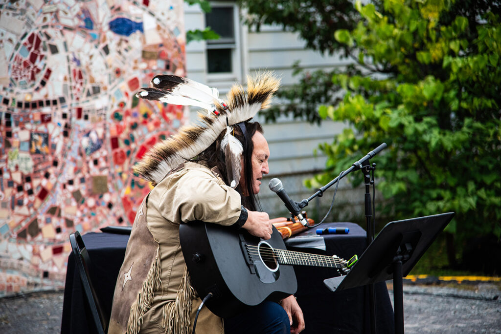 Bill Miller sits with his guitar on the Freedom Square stage, wearing his traditional Mohican garb, including a feather headdress