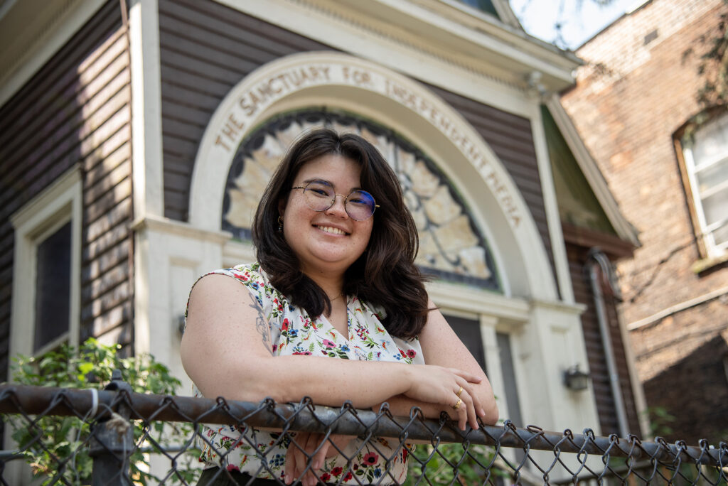 Ren Lee, an Asian-American person with long, brown hair and circle glasses, leans on the chain linked fence with the Sanctuary building towering behind them. 