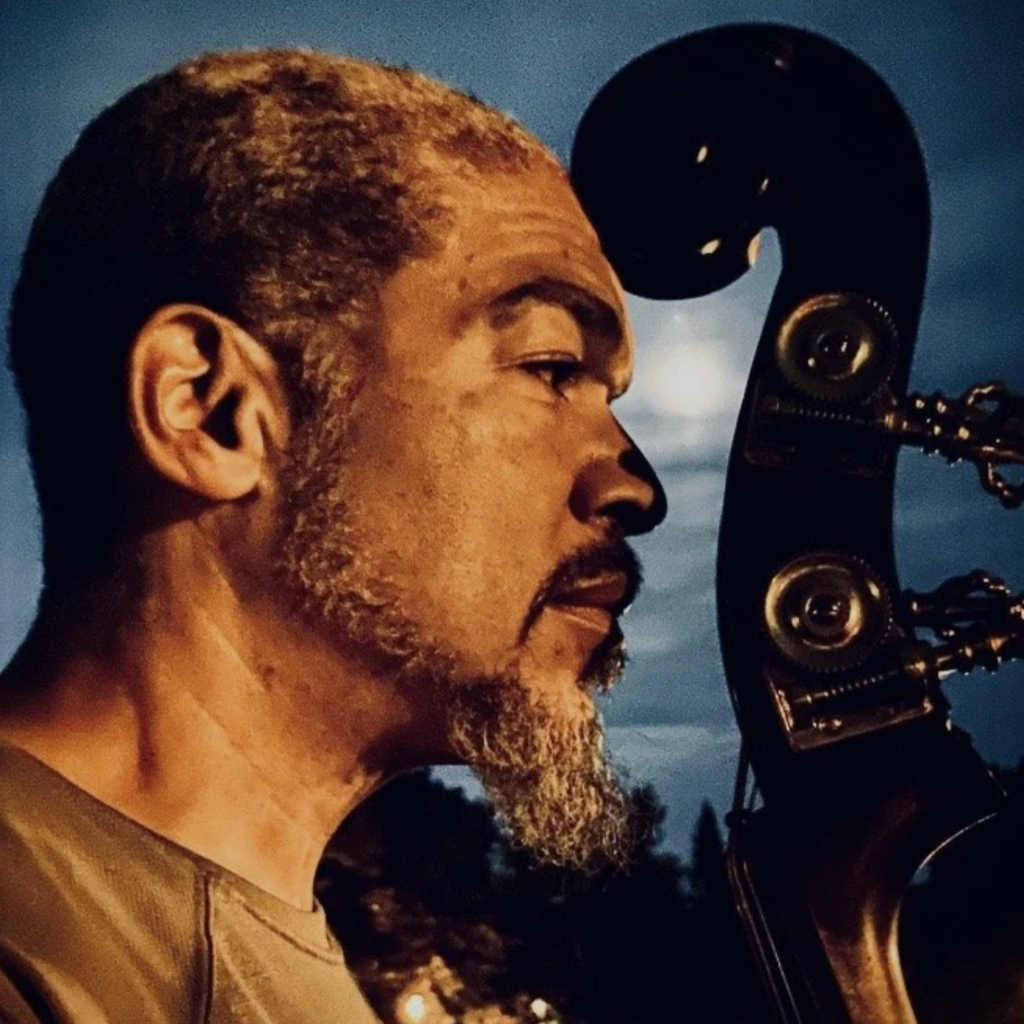 A profile picture of Tarik Shah in evening light with Tarik's face very close to his bass's headstock, the curved wood at the top of the instrument
