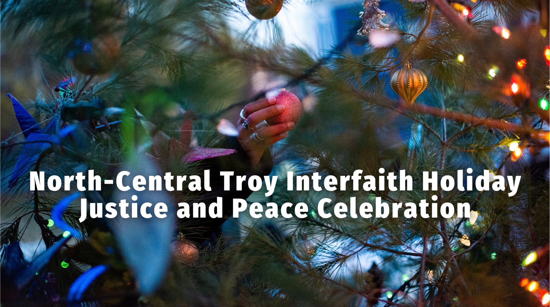 a had puts a holiday ornament in the evergreen tree. white text says, north-central troy interfaith holiday justice and peace celebration