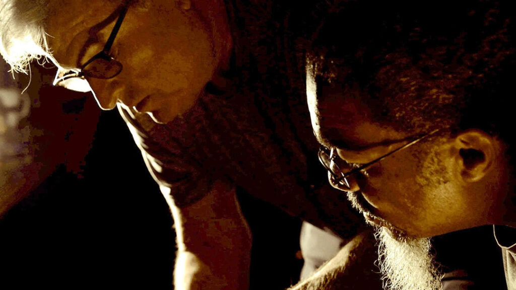A close up photo of Barry Rubinow and Tarik Shah bending over, just slightly illuminated by light.