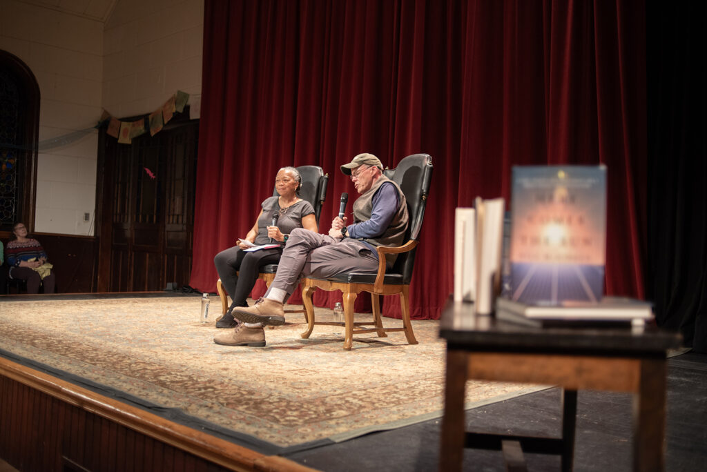 Bill McKibben sits on stage with Benita Law-Diao. At the right of the frame is a stack of McKibben's books "Here Comes the Sun"