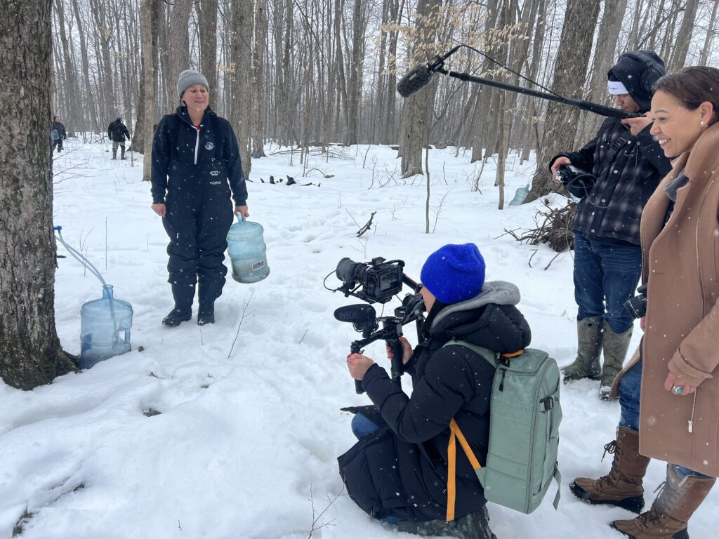 An indigenous woman collecting maple sap in large plastic jugs in a snowy forest is being filmed by a film crew of indigenous women