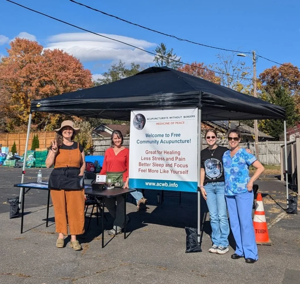 a pop up tent with a sign saying "welcome to free community acupuncture" next to which four people are standing. Gina Mauro is standing at the far left holding up a peace sign with her finders and wearing a brimmed sun hat. 