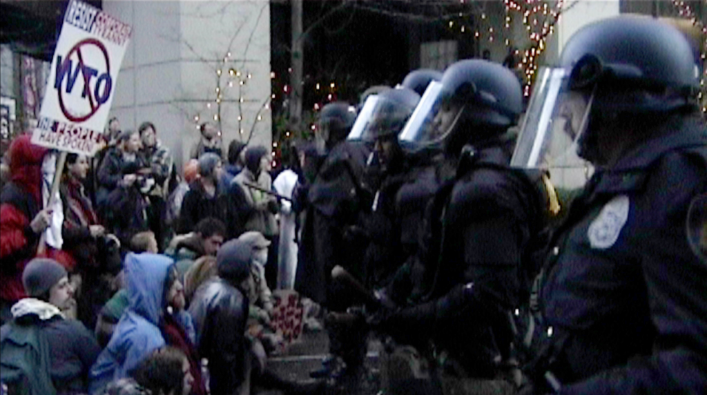 a film still from the film "WTO/99" showing a line of swat police at the right facing a group of civilians to the left, one person holds a sign of WTO in a slashed circle