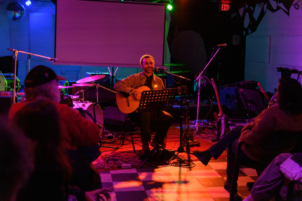 Abu Zeyn smiling while holding his guitar, seated in front of an audience