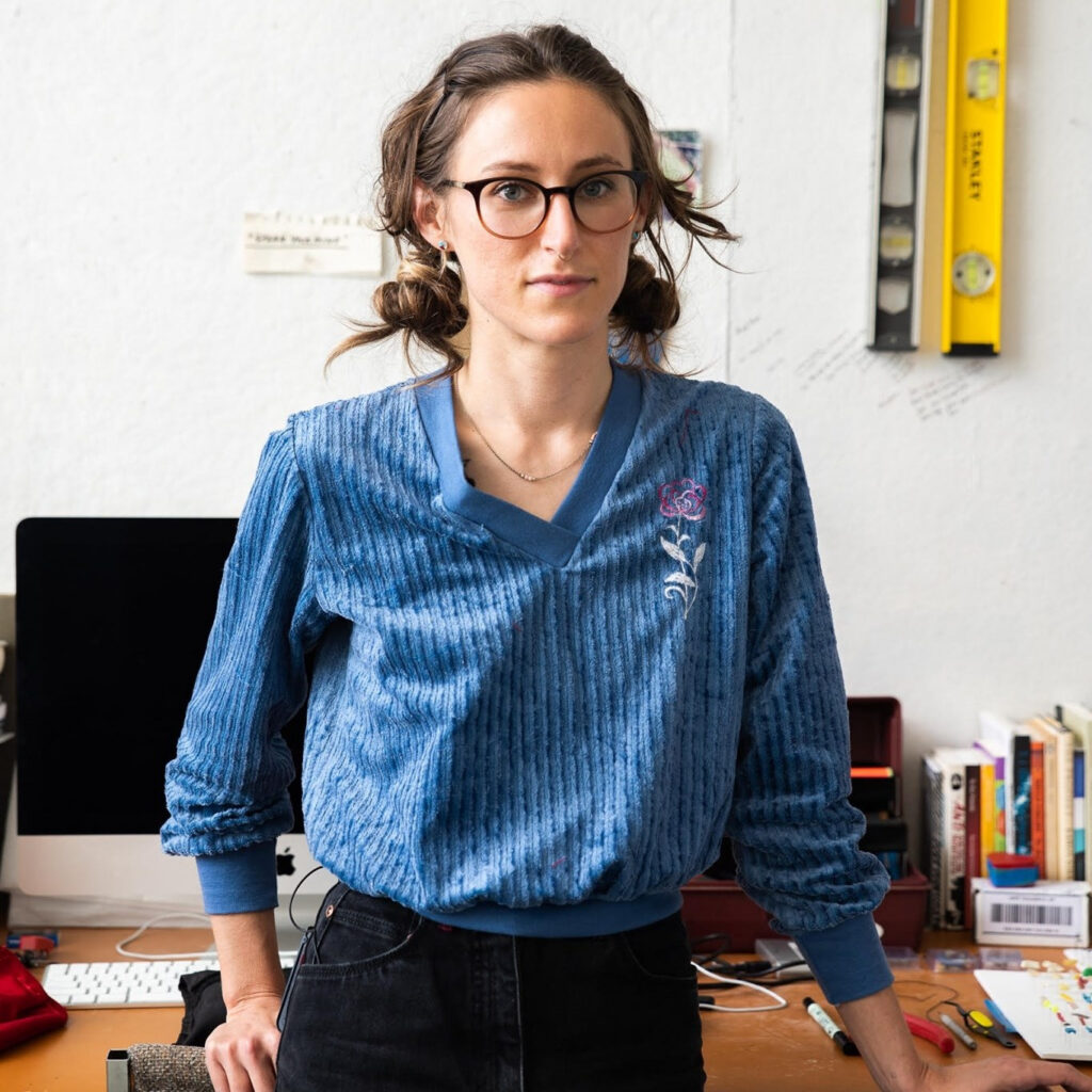 A person wearing glasses with brown hair up in two low buns and wearing a baggy blue shirt looks into the camera.